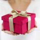 Close-up of hands offering a crimson gift box with a gold ribbon.