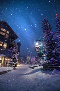 Snowy village street at night, decorated for Christmas. Festive lights illuminate snow-covered trees and buildings.